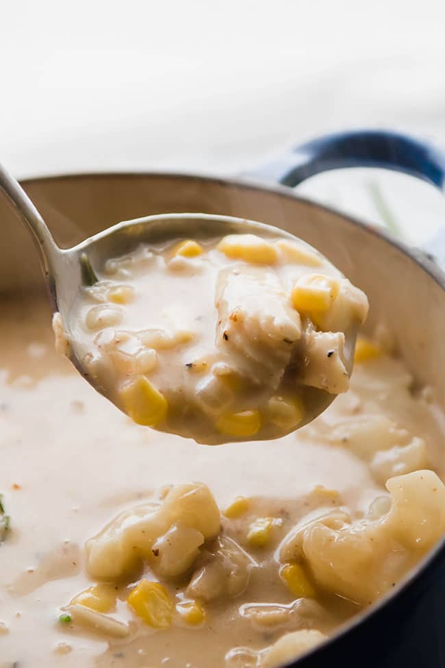 Silver ladle lifting a scoop of fish and corn chowder out of a large pot.