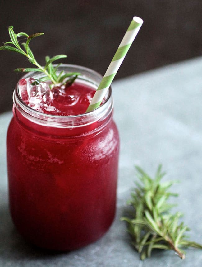 Pink cocktail in a glass mason jar on a white tablecloth, topped with a sprig of fresh rosemary and a green paper straw.