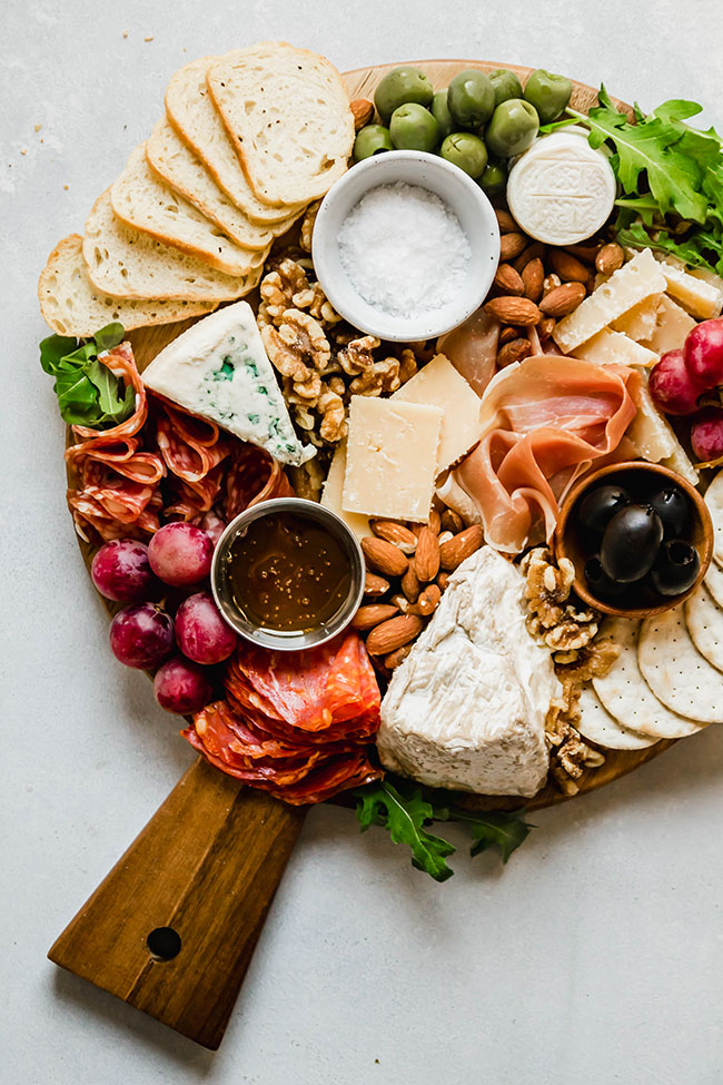 Cheese and charcuterie on a round wooden cutting board on a white table.