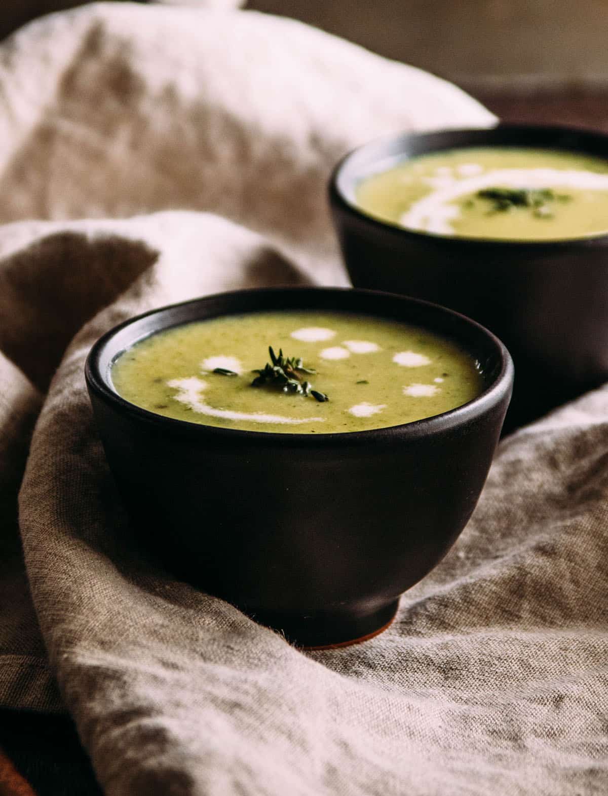 Green soup in a small black bowl, next to a a light brown linen napkin.