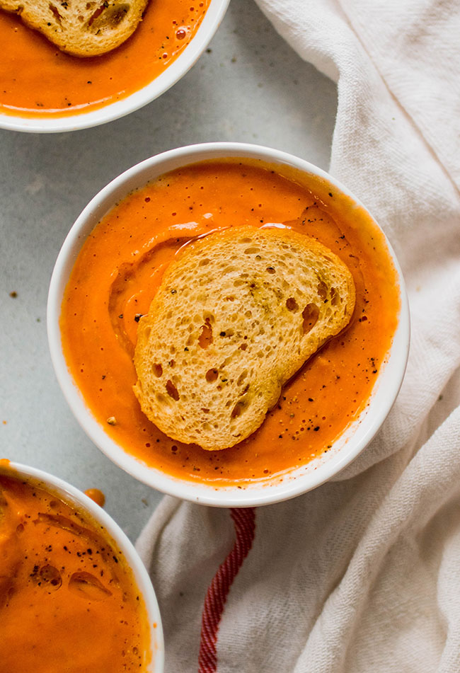 Creamy tomato soup in a white bowl topped with a toasted baguette slice.