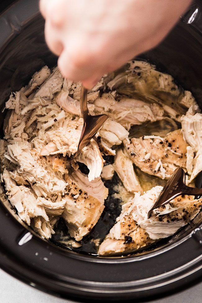 Man\'s hands using forks to shred cooked chicken in the bowl of a slow cooker.