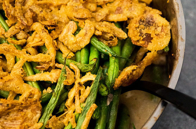 Spoon lifting green beans topped with fried onions out of a baking dish.