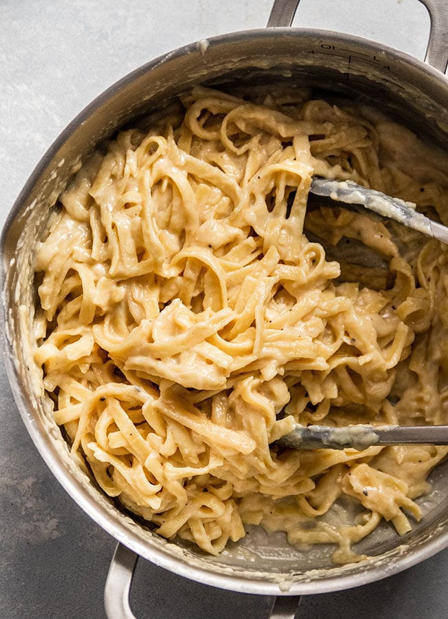 Tongs mixing fettuccine alfredo in a silver pot.