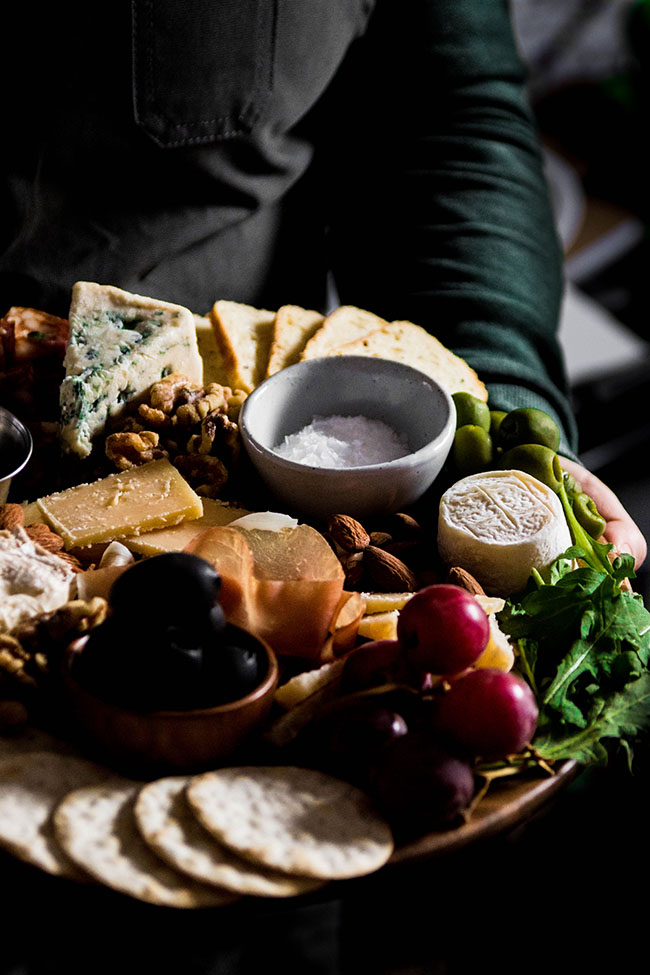Woman in a green shirt holding a cheese board with goat cheese, grapes, black olives, and blue cheese.