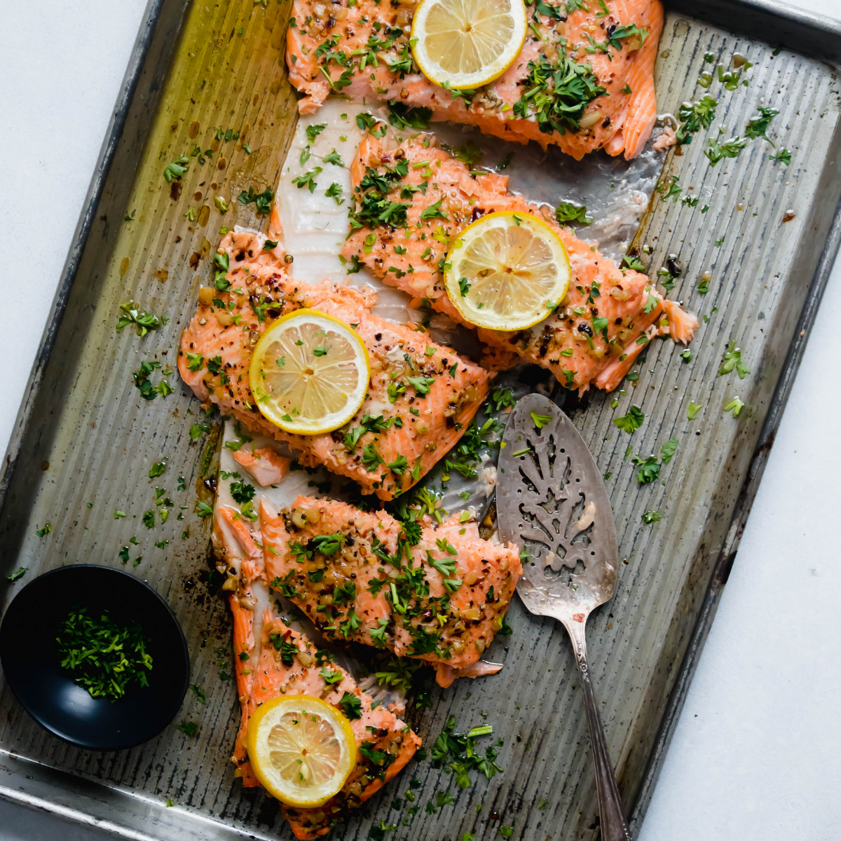 Baked steelhead fillet on a metal sheet pan, topped with fresh parsley and lemon slices and cut into portions.