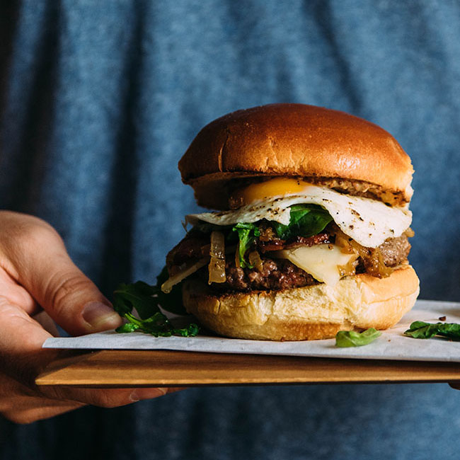 Man in a blue shirt holding a wood cutting board with a cheeseburger on top.