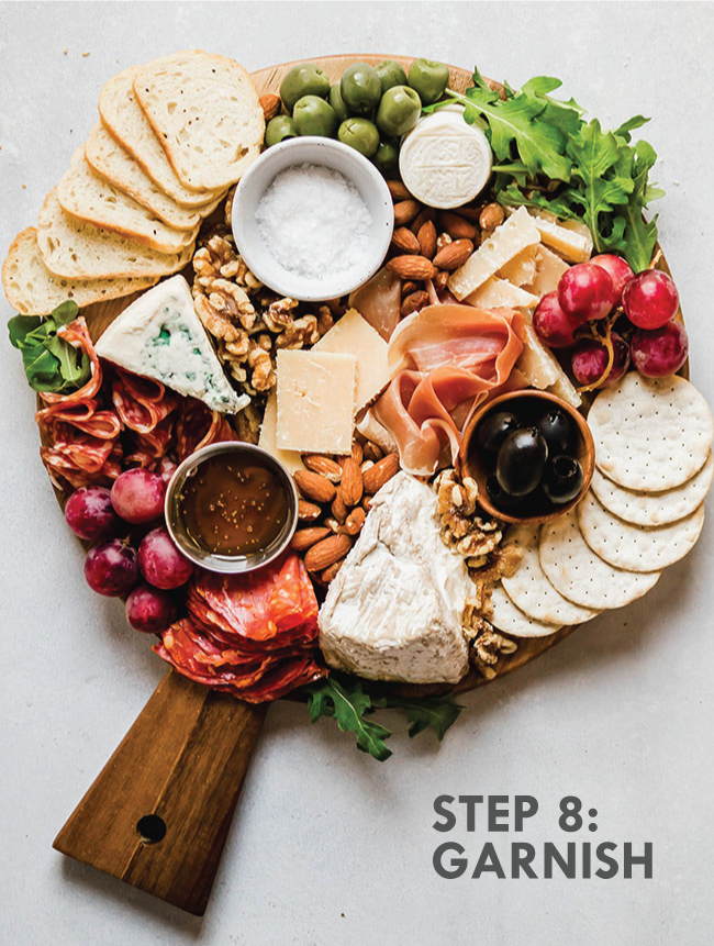 A completed cheese plate with fresh arugula tucked around the edges. Grey text overlay says \'step 8: garnish.\'