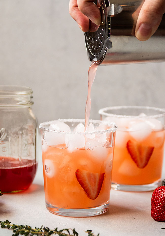 Man's hand pouring strawberry margarita into a glass with a metal cocktail shaker.