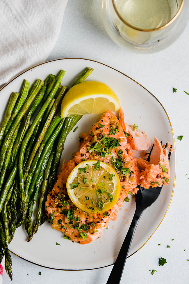 Roasted steelhead topped with lemon slice and parsley on a white plate with roasted asparagus.