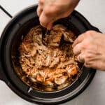 Man's hands shredding chicken in a black crock pot.