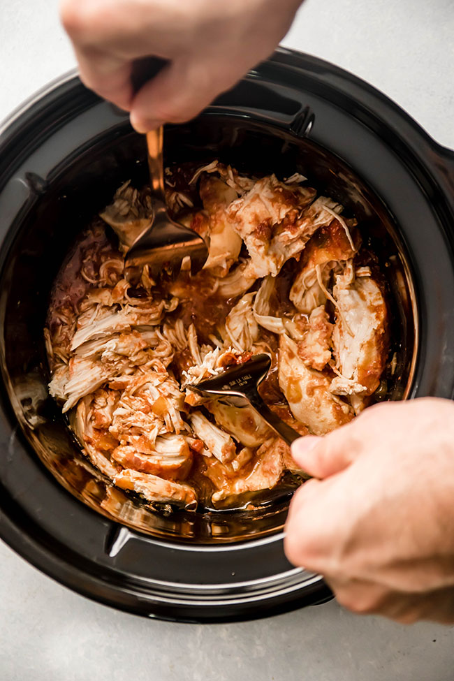 Hands using two forks to shred chicken in a black bowl.