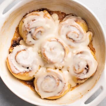 Six cinnamon rolls with cream cheese frosting in a white baking dish.