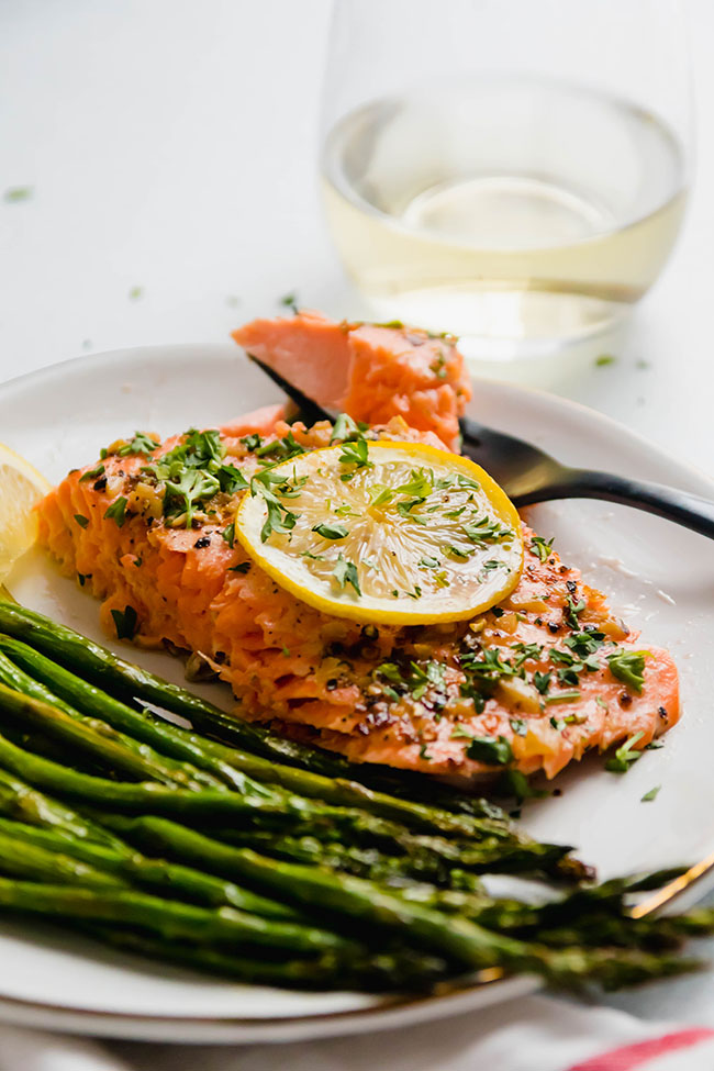 Slice of baked steelhead topped with a lemon slice on a white plate next to roasted asparagus.