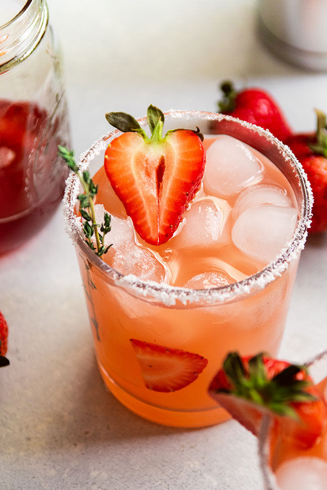 Strawberry margarita on a white table, garnished with a sprig of thyme and half of a fresh strawberry.