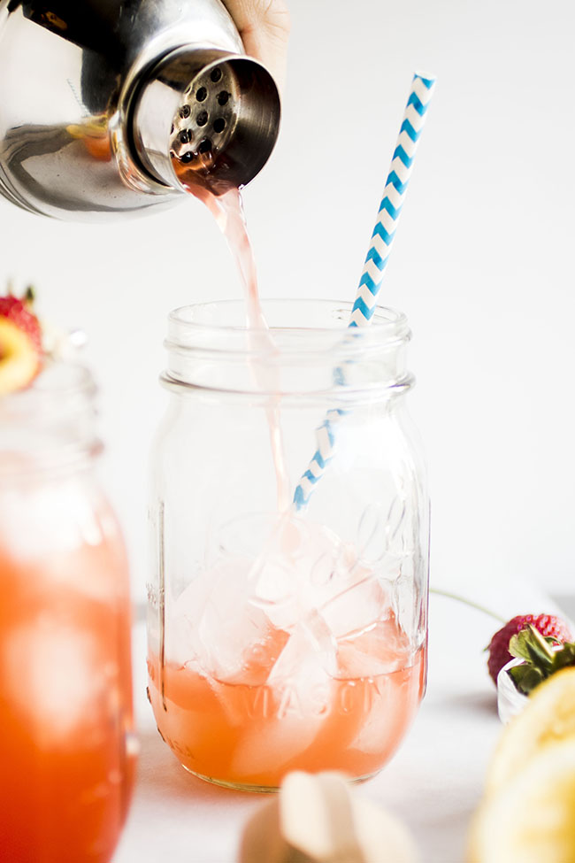 Silver cocktail shaker pouring cocktail into mason jar with a blue straw.