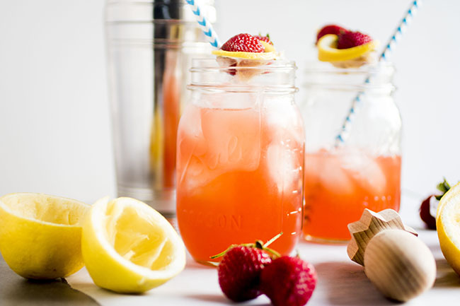 Two mason jars of strawberry cocktail, next to fresh strawberries, a lemon, and several cocktail tools.