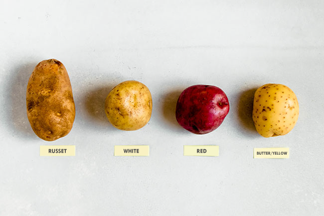 Four potatoes, russet, white, red, and butter or yellow, arranged in a row on a white surface.