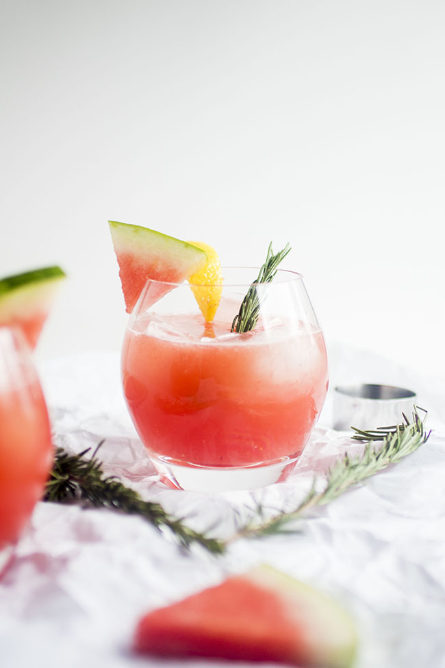 Watermelon cocktail on a white table next to several sprigs of fresh rosemary.