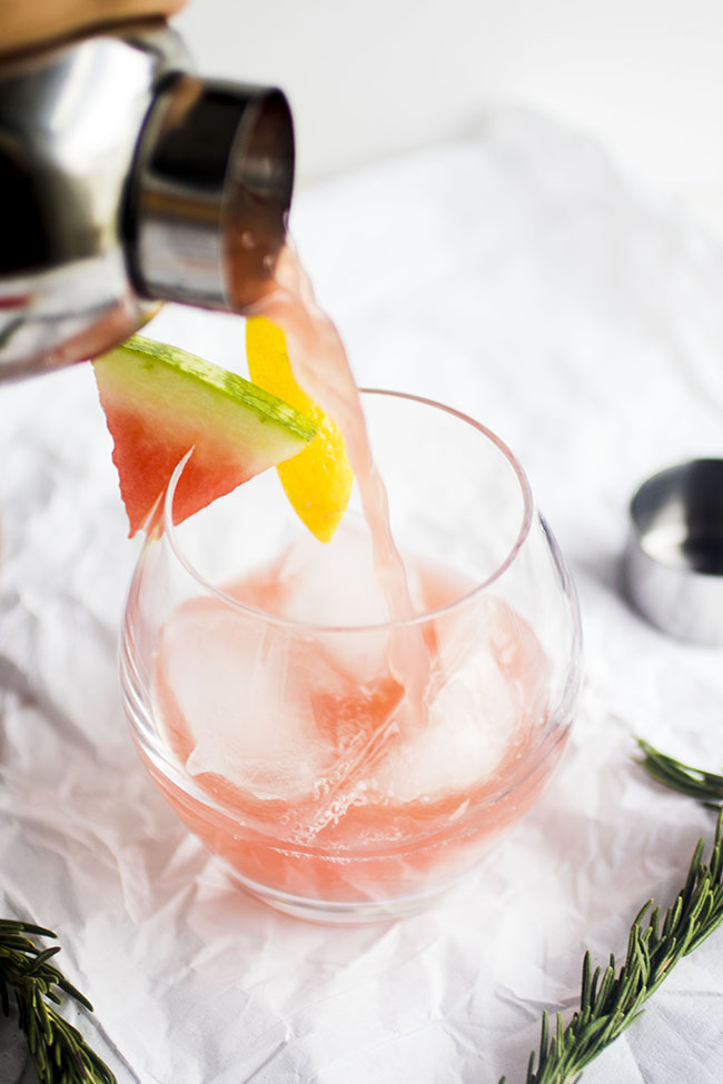 Silver cocktail shaker pouring watermelon cocktail into a short glass.