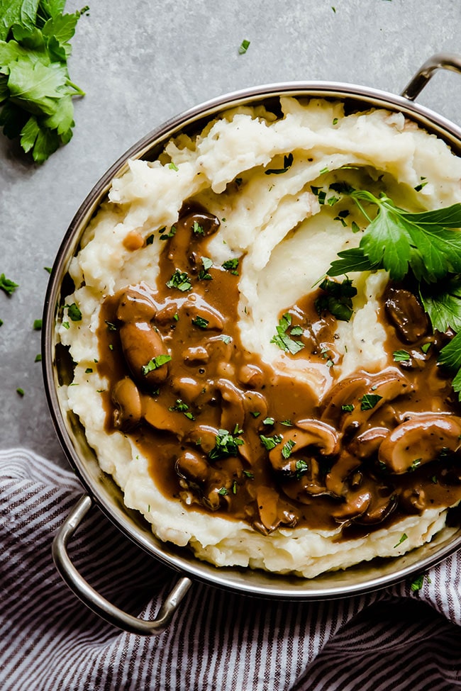Mashed potatoes topped with mushroom gravy in a silver serving dish on a grey table.